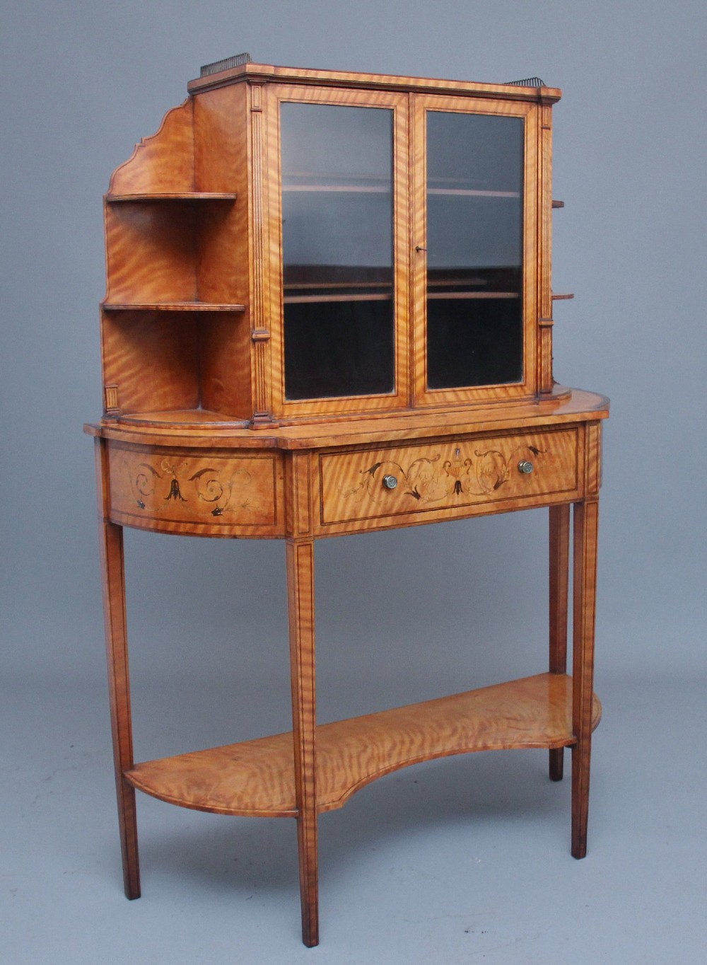A 19th-century satinwood display cabinet featuring central glazed double doors, curved side shelves, a marquetry-inlaid drawer, and slender tapered legs with a lower undertier. View XII