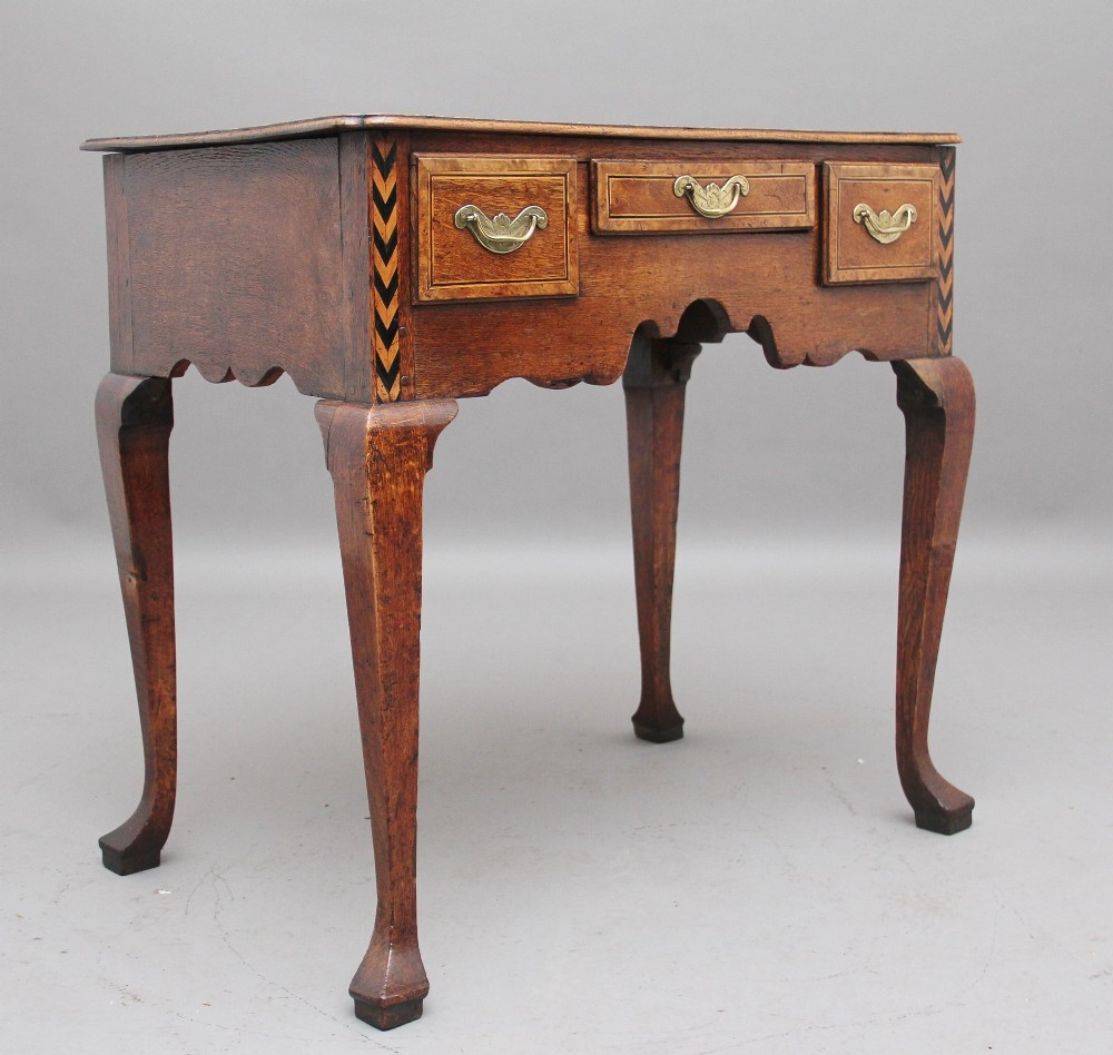 An 18th-century oak lowboy table featuring three drawers with brass handles, inlaid chevron borders, a scalloped apron, and four cabriole legs.