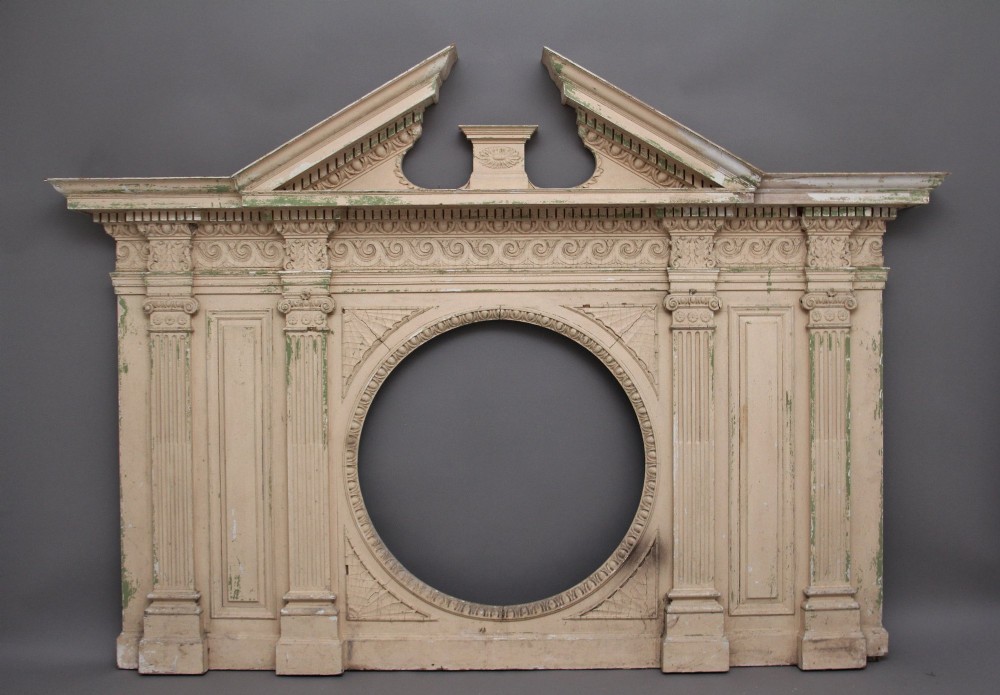 An 18th-century carved pine architectural overmantel featuring a broken pediment, fluted pilasters, dentil mouldings, and a central circular aperture in distressed off-white paint.