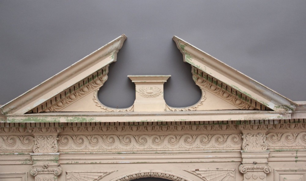 An 18th-century carved pine architectural overmantel featuring a broken pediment, fluted pilasters, dentil mouldings, and a central circular aperture in distressed off-white paint. View II