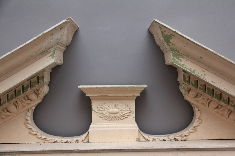 An 18th-century carved pine architectural overmantel featuring a broken pediment, fluted pilasters, dentil mouldings, and a central circular aperture in distressed off-white paint. View III