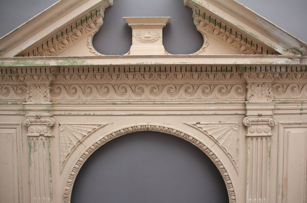 An 18th-century carved pine architectural overmantel featuring a broken pediment, fluted pilasters, dentil mouldings, and a central circular aperture in distressed off-white paint. View VI