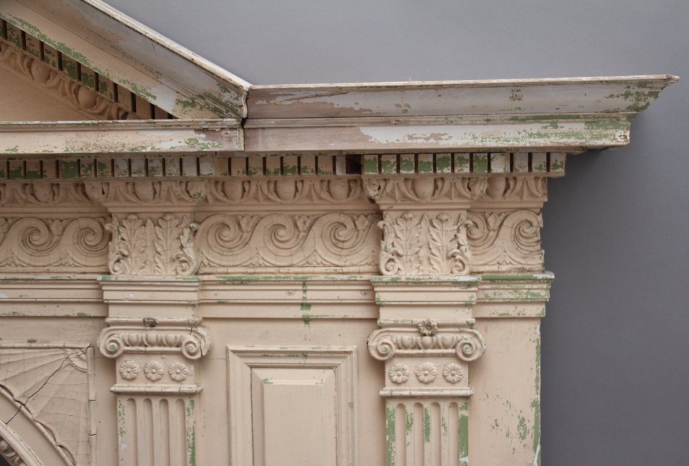 An 18th-century carved pine architectural overmantel featuring a broken pediment, fluted pilasters, dentil mouldings, and a central circular aperture in distressed off-white paint. View VII