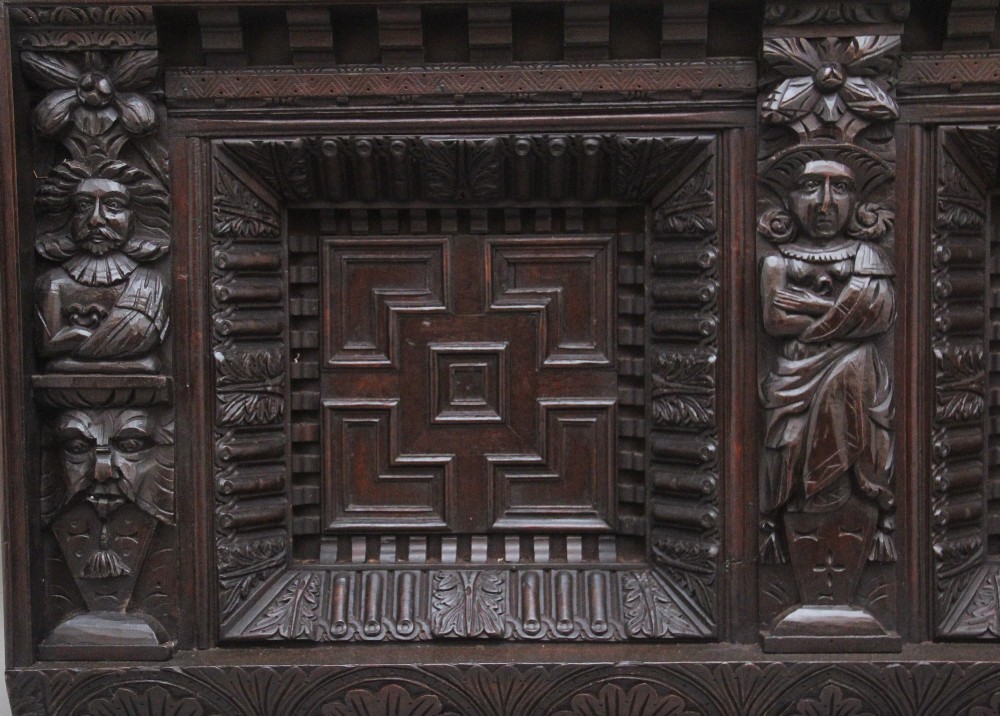 A rectangular dark carved oak overmantel panel with geometric recessed centre panels flanked by carved human figures and mask motifs. View II