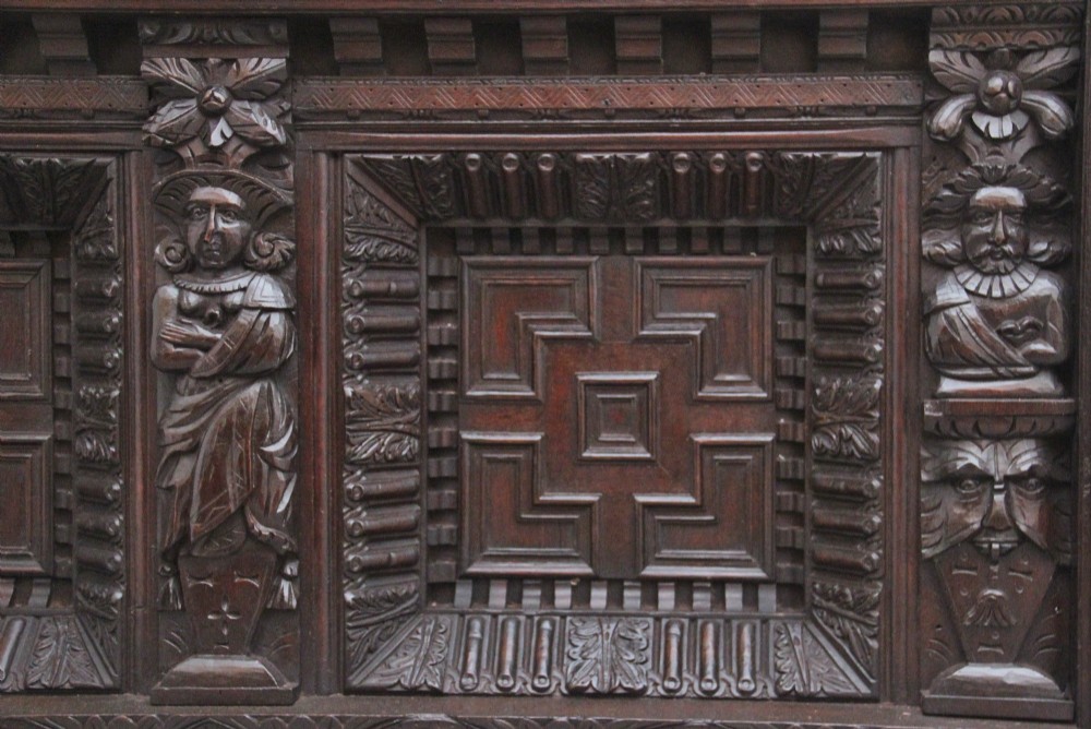 A rectangular dark carved oak overmantel panel with geometric recessed centre panels flanked by carved human figures and mask motifs. View III