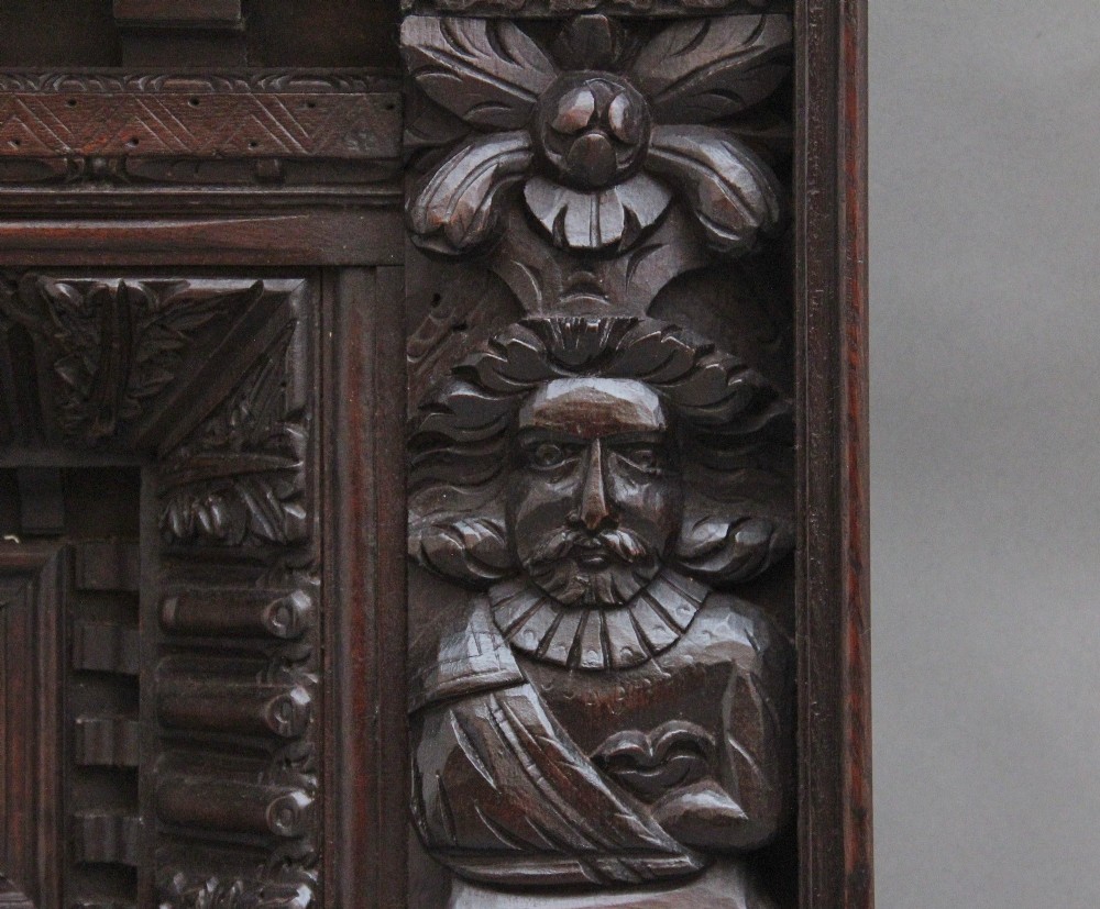A rectangular dark carved oak overmantel panel with geometric recessed centre panels flanked by carved human figures and mask motifs. View IV