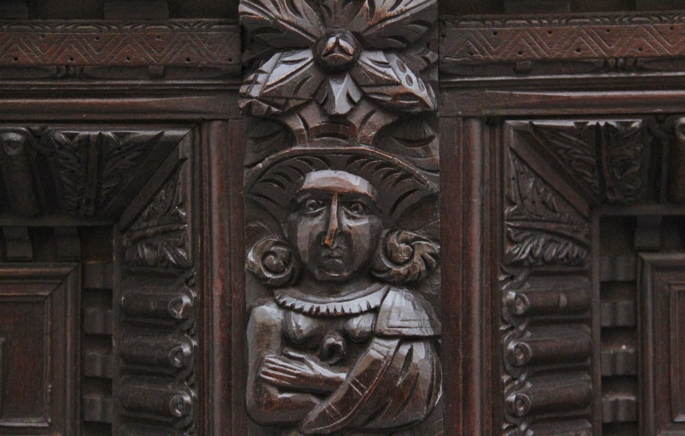 A rectangular dark carved oak overmantel panel with geometric recessed centre panels flanked by carved human figures and mask motifs. View V