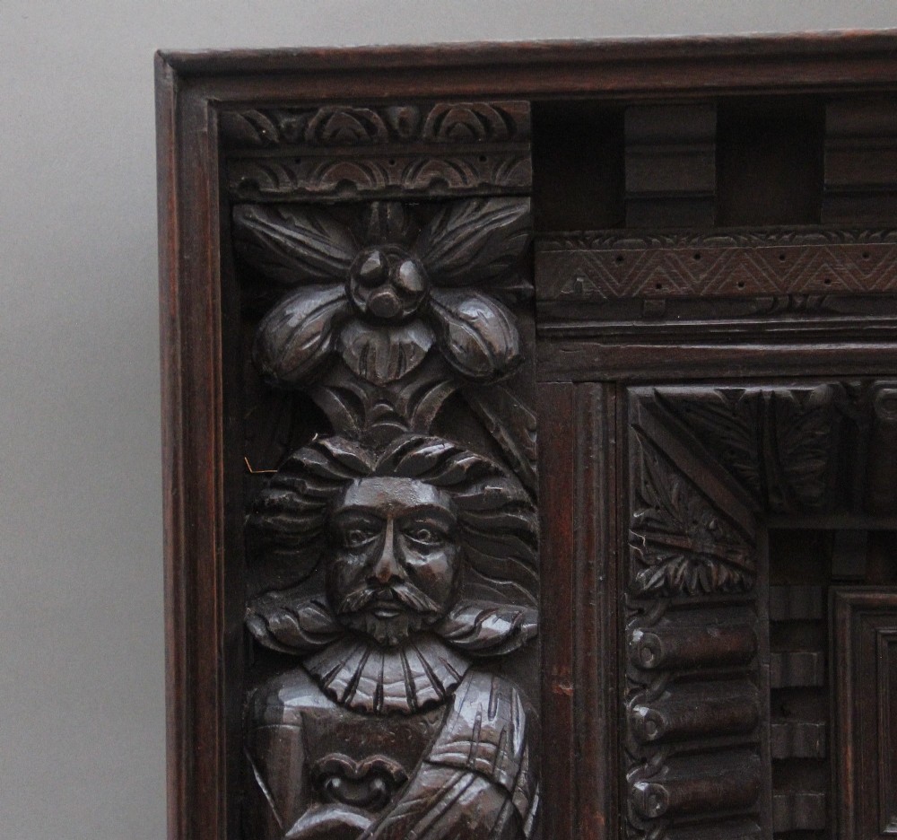 A rectangular dark carved oak overmantel panel with geometric recessed centre panels flanked by carved human figures and mask motifs. View VI