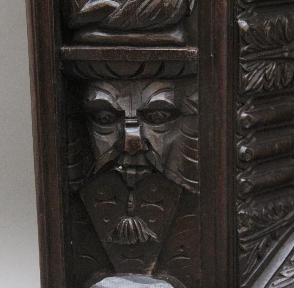 A rectangular dark carved oak overmantel panel with geometric recessed centre panels flanked by carved human figures and mask motifs. View VII
