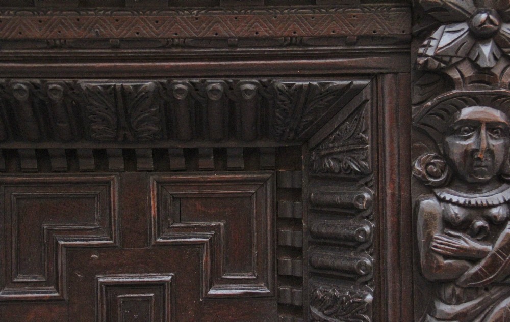 A rectangular dark carved oak overmantel panel with geometric recessed centre panels flanked by carved human figures and mask motifs. View VIII