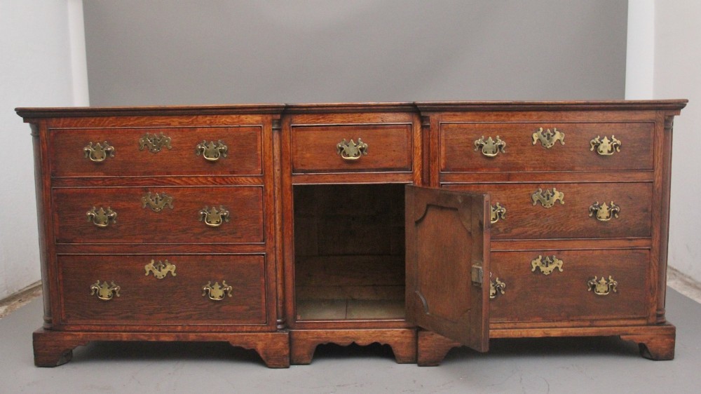 A large 18th-century oak dresser base featuring brass swan-neck handles, multiple drawers, a central panelled cupboard, and bracket feet. View II