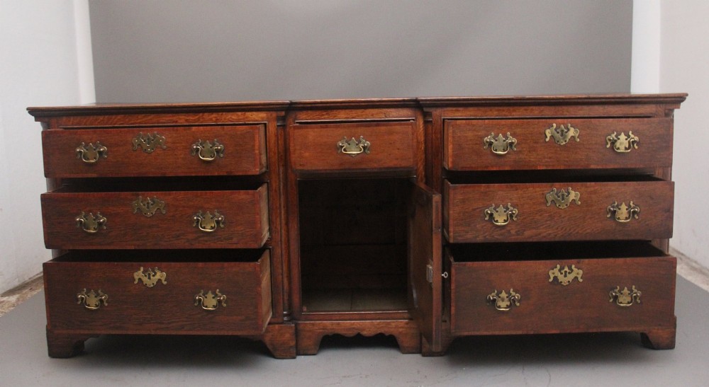 A large 18th-century oak dresser base featuring brass swan-neck handles, multiple drawers, a central panelled cupboard, and bracket feet. View III