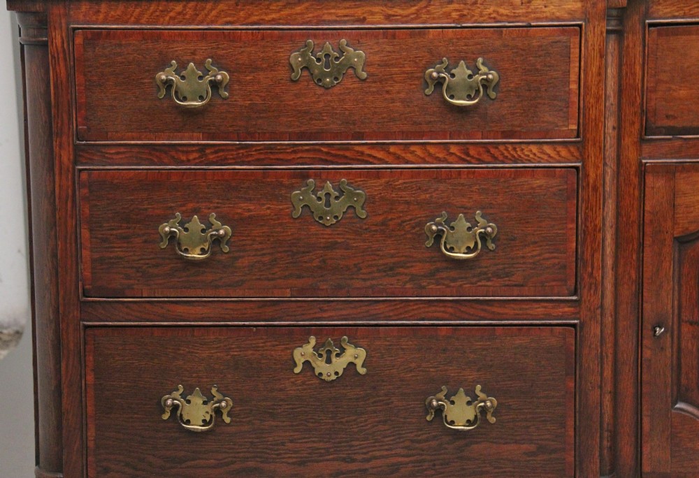 A large 18th-century oak dresser base featuring brass swan-neck handles, multiple drawers, a central panelled cupboard, and bracket feet. View V