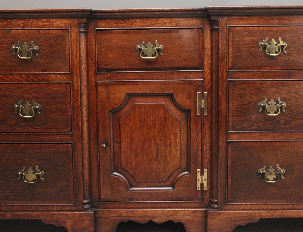 A large 18th-century oak dresser base featuring brass swan-neck handles, multiple drawers, a central panelled cupboard, and bracket feet. View VI