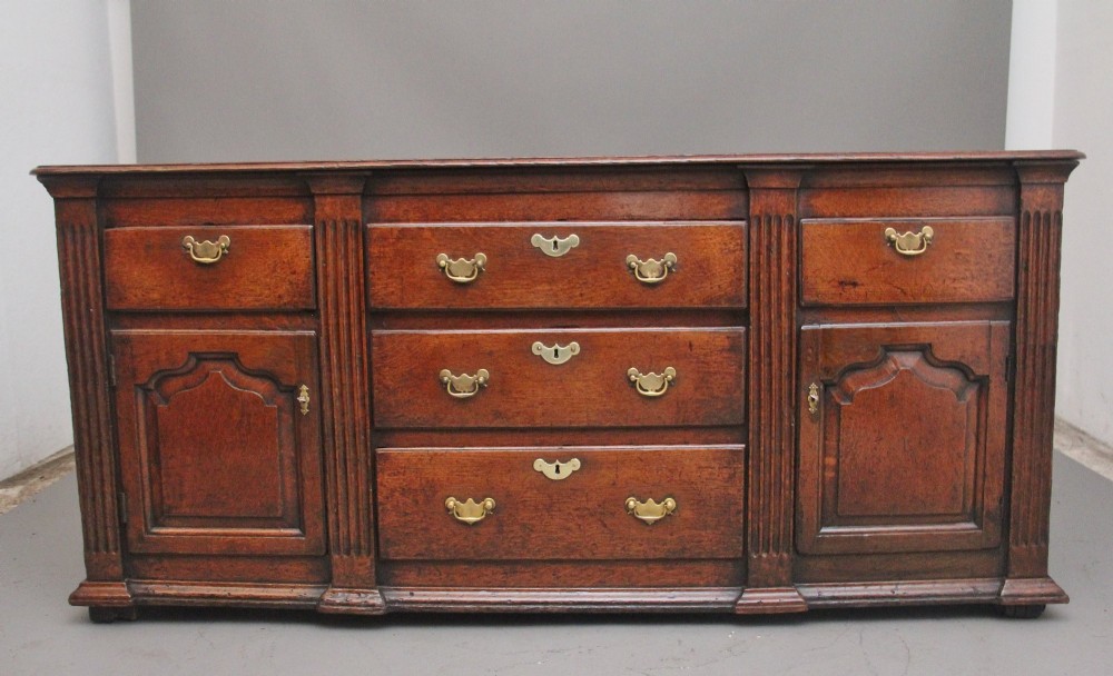 A large 18th-century oak dresser base featuring brass swan-neck handles, multiple drawers, a central panelled cupboard, and bracket feet. View XII