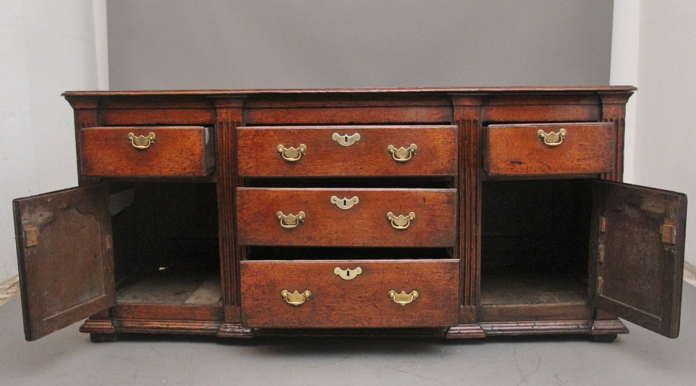 A large 18th-century oak dresser base featuring brass swan-neck handles, multiple drawers, a central panelled cupboard, and bracket feet. View XIII