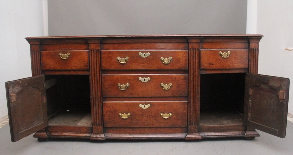 A large 18th-century oak dresser base featuring brass swan-neck handles, multiple drawers, a central panelled cupboard, and bracket feet. View XIV