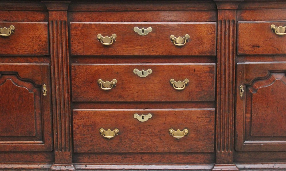 A large 18th-century oak dresser base featuring brass swan-neck handles, multiple drawers, a central panelled cupboard, and bracket feet. View XV