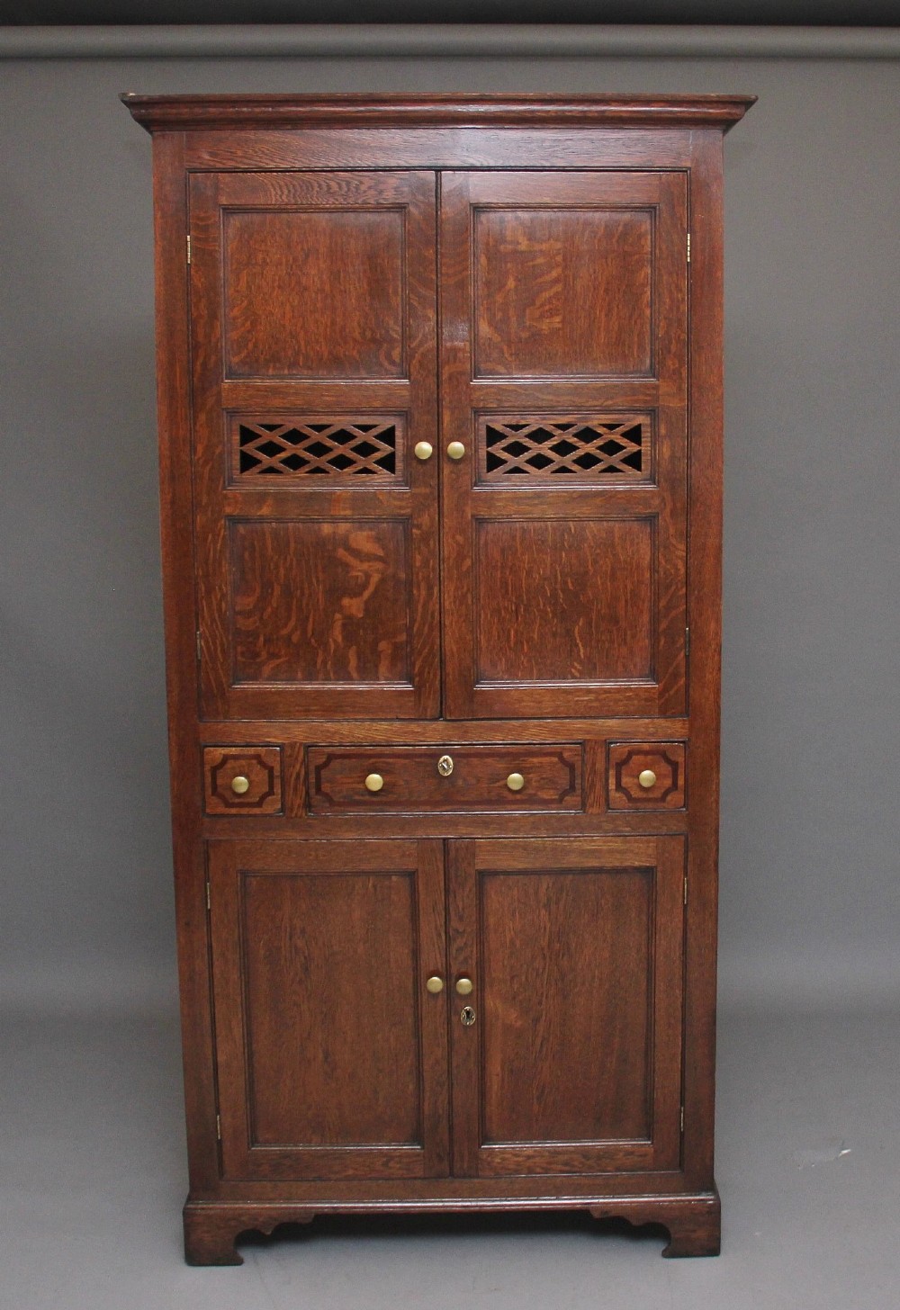 An early 19th-century dark oak food cupboard featuring pierced lattice ventilation panels, brass drawer handles, and panelled double doors.