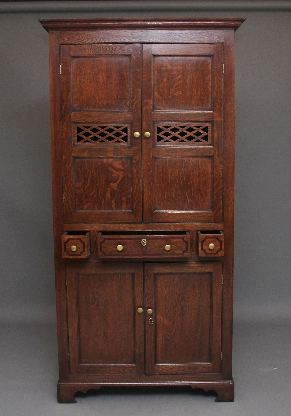 An early 19th-century dark oak food cupboard featuring pierced lattice ventilation panels, brass drawer handles, and panelled double doors. View III