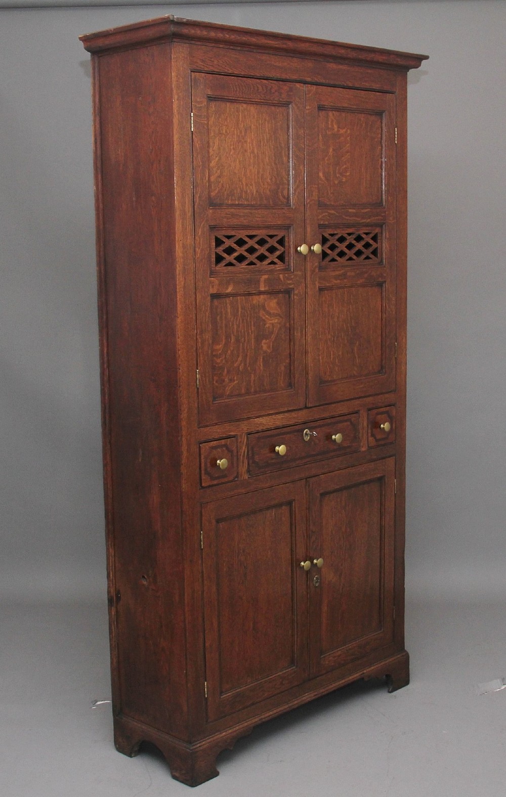 An early 19th-century dark oak food cupboard featuring pierced lattice ventilation panels, brass drawer handles, and panelled double doors. View IV