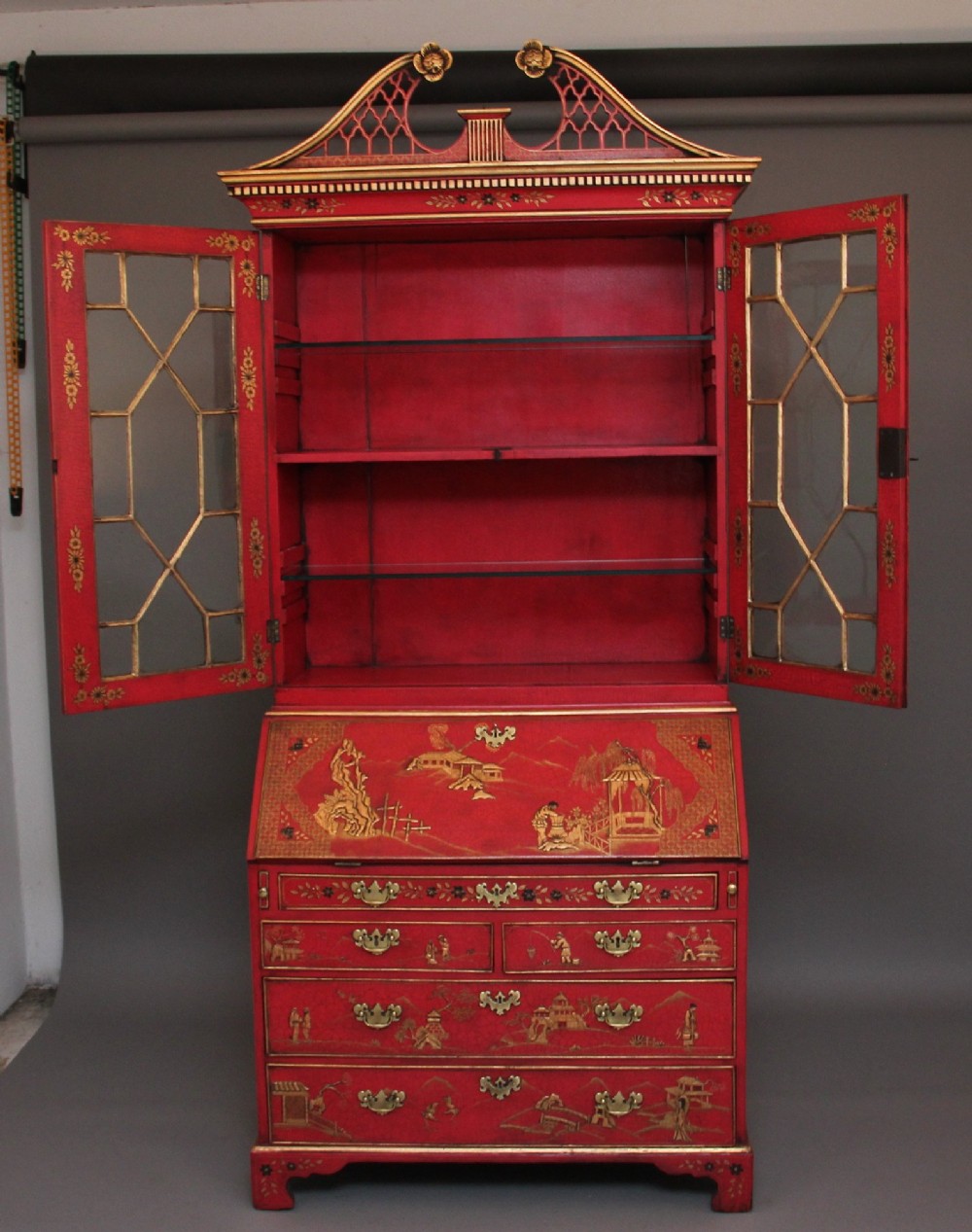 An early twentieth-century red lacquered Chinoiserie bureau bookcase decorated with gilded landscapes, double glazed doors, and brass drop handles. View II