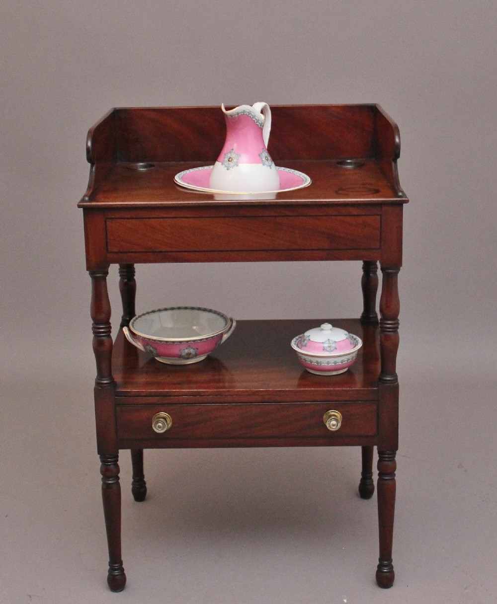 An early Victorian mahogany washstand featuring a gallery splashback top with circular aperture cutouts, a central open shelf, turned legs, and a bottom drawer with brass handles.