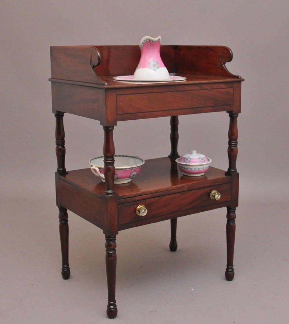 An early Victorian mahogany washstand featuring a gallery splashback top with circular aperture cutouts, a central open shelf, turned legs, and a bottom drawer with brass handles. View IV