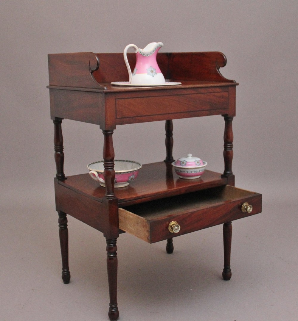 An early Victorian mahogany washstand featuring a gallery splashback top with circular aperture cutouts, a central open shelf, turned legs, and a bottom drawer with brass handles. View V