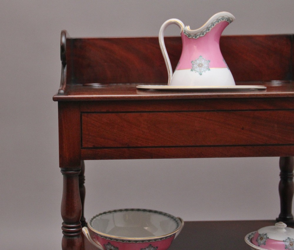An early Victorian mahogany washstand featuring a gallery splashback top with circular aperture cutouts, a central open shelf, turned legs, and a bottom drawer with brass handles. View VI