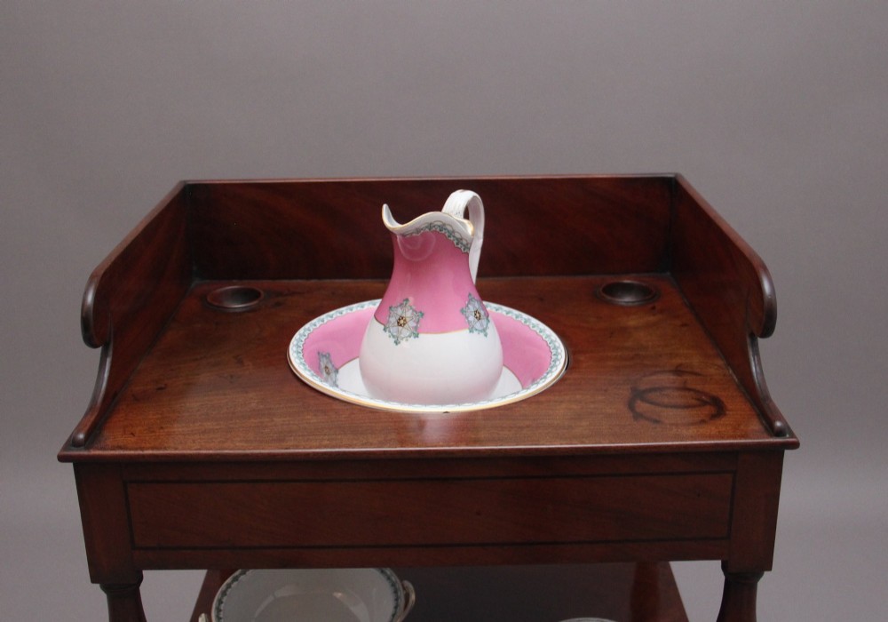 An early Victorian mahogany washstand featuring a gallery splashback top with circular aperture cutouts, a central open shelf, turned legs, and a bottom drawer with brass handles. View VII