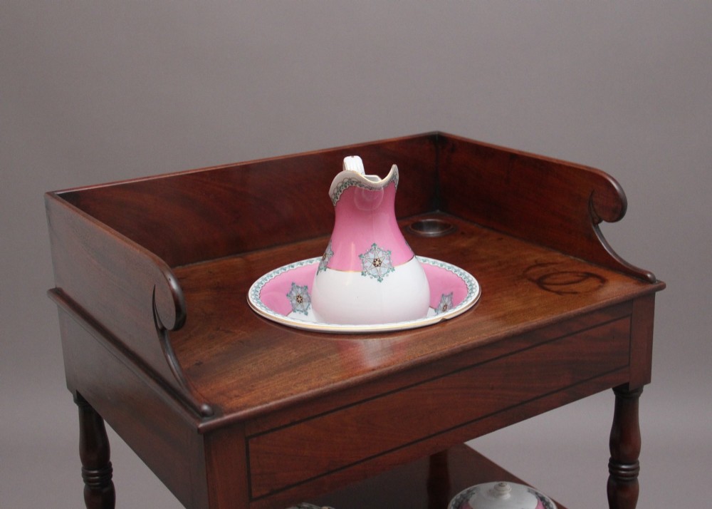 An early Victorian mahogany washstand featuring a gallery splashback top with circular aperture cutouts, a central open shelf, turned legs, and a bottom drawer with brass handles. View IX