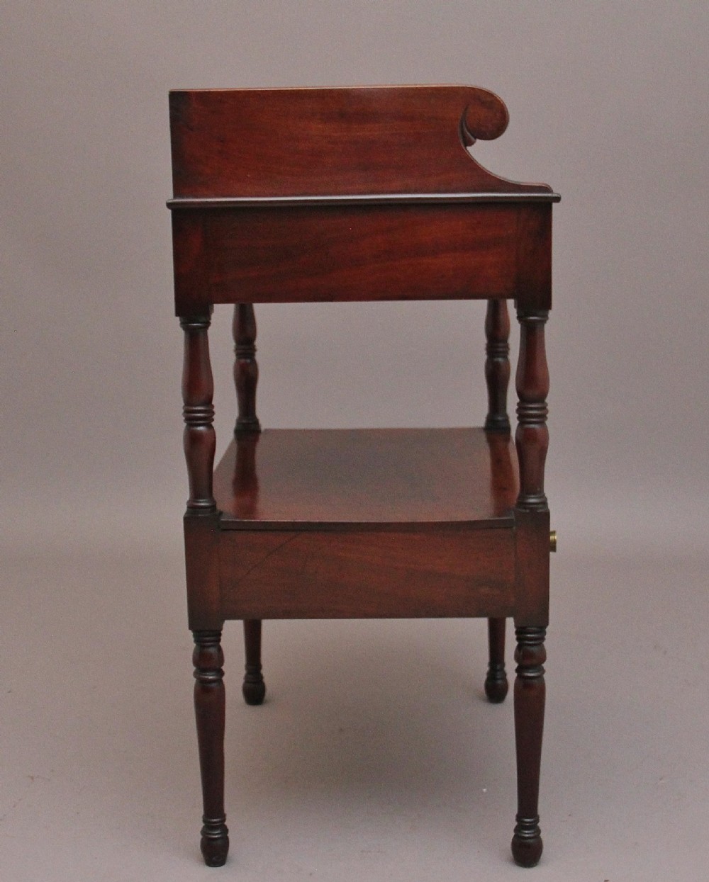 An early Victorian mahogany washstand featuring a gallery splashback top with circular aperture cutouts, a central open shelf, turned legs, and a bottom drawer with brass handles. View XII