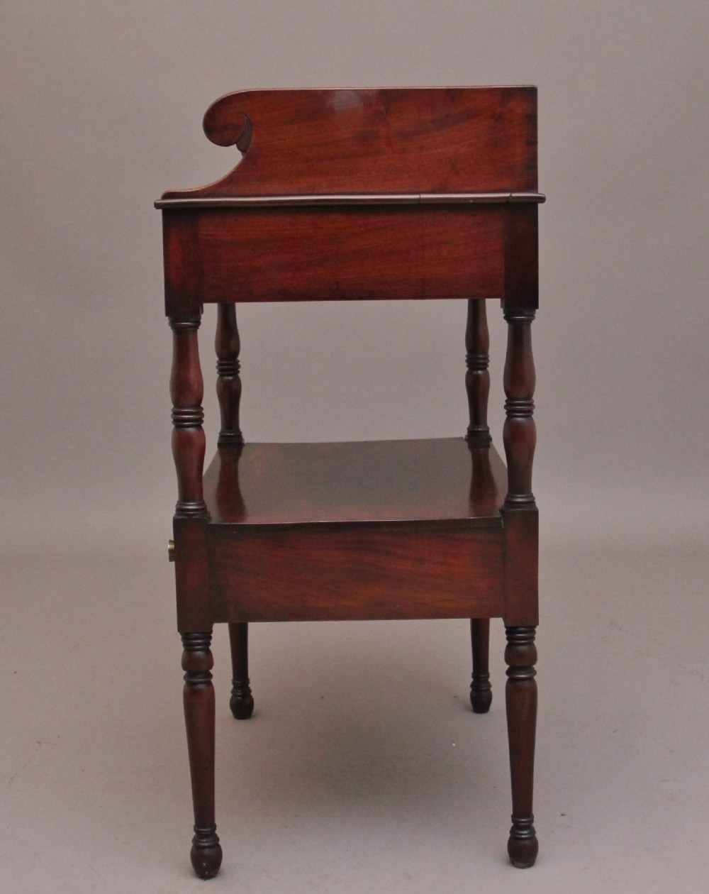 An early Victorian mahogany washstand featuring a gallery splashback top with circular aperture cutouts, a central open shelf, turned legs, and a bottom drawer with brass handles. View XIV