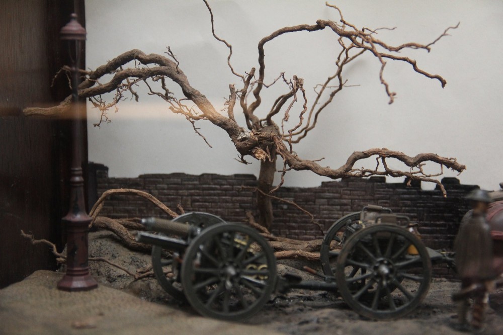 A glazed wooden display case containing an early 20th-century military diorama of a horse-drawn artillery team on a WWI battlefield scene. View IV