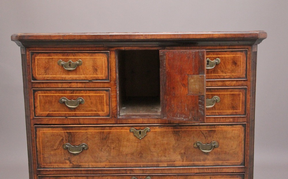 An early 18th-century George II walnut chest of drawers featuring crossbanded drawer fronts, brass handles, a central cupboard door, and bracket feet. View III