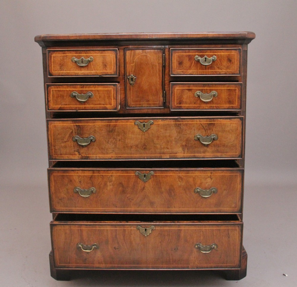An early 18th-century George II walnut chest of drawers featuring crossbanded drawer fronts, brass handles, a central cupboard door, and bracket feet. View V