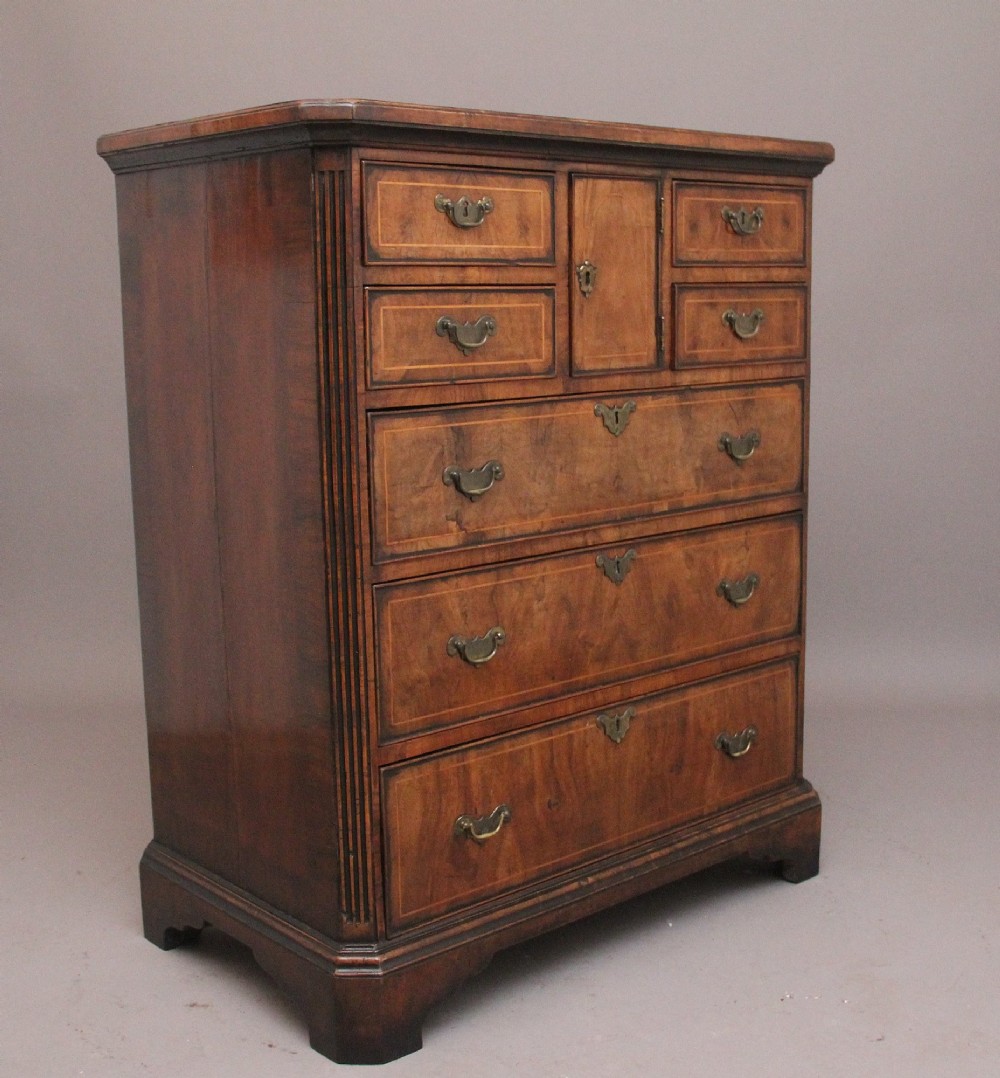 An early 18th-century George II walnut chest of drawers featuring crossbanded drawer fronts, brass handles, a central cupboard door, and bracket feet. View VI
