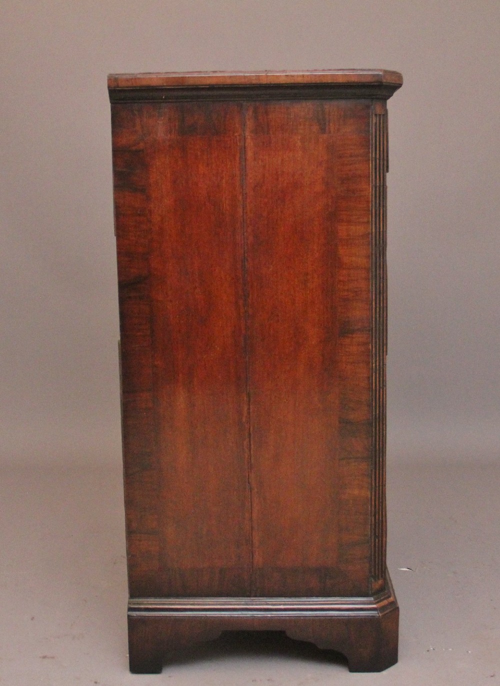 An early 18th-century George II walnut chest of drawers featuring crossbanded drawer fronts, brass handles, a central cupboard door, and bracket feet. View VIII