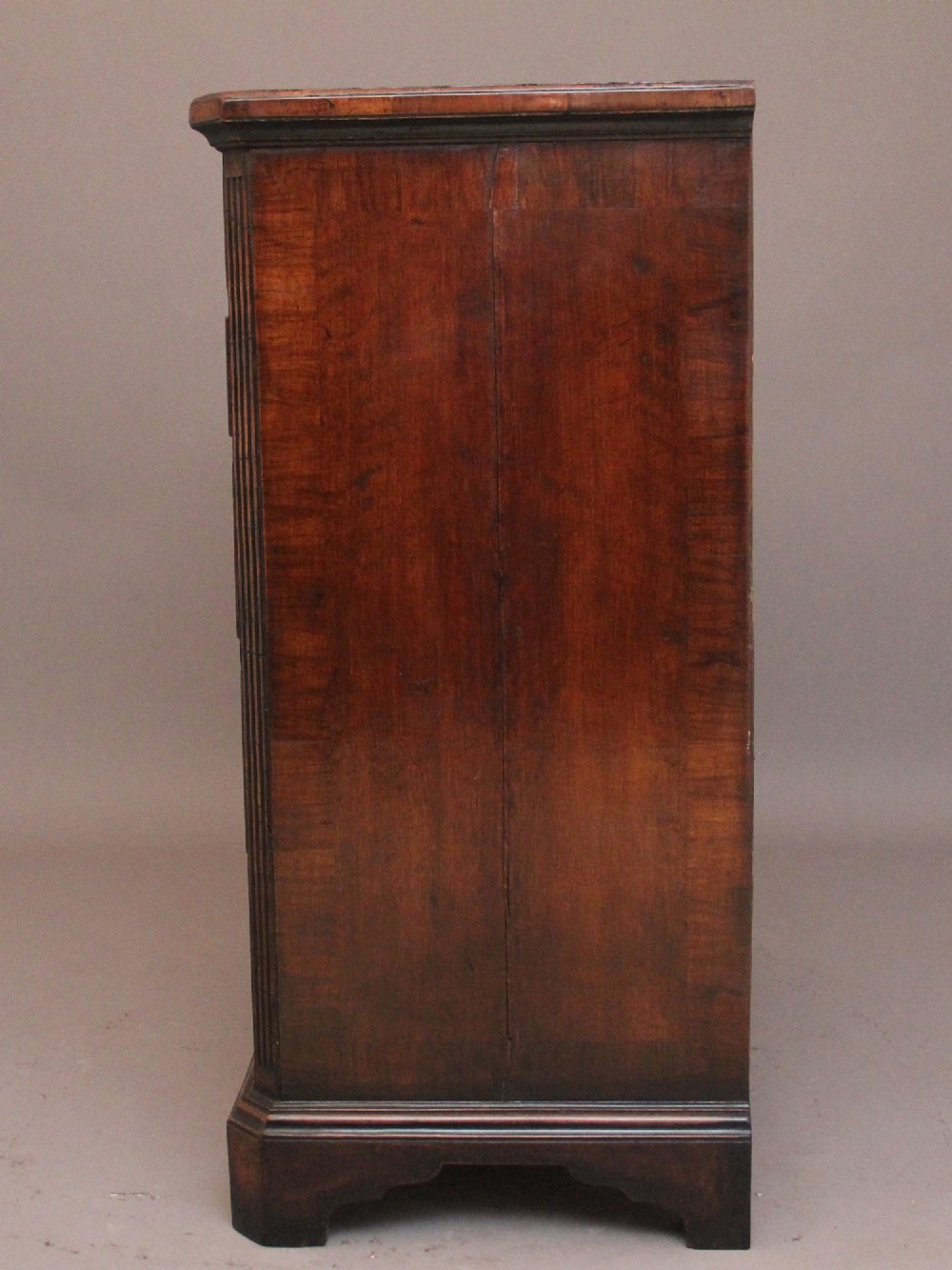 An early 18th-century George II walnut chest of drawers featuring crossbanded drawer fronts, brass handles, a central cupboard door, and bracket feet. View X
