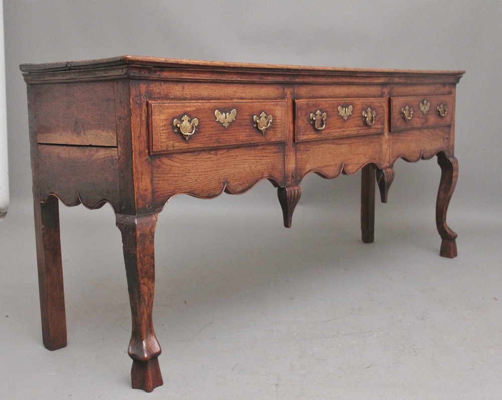 An early 18th-century oak dresser base featuring three brass-handled drawers, a scalloped apron with pendant drops, and cabriole legs. View VI