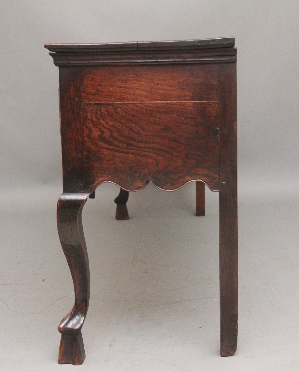 An early 18th-century oak dresser base featuring three brass-handled drawers, a scalloped apron with pendant drops, and cabriole legs. View IX