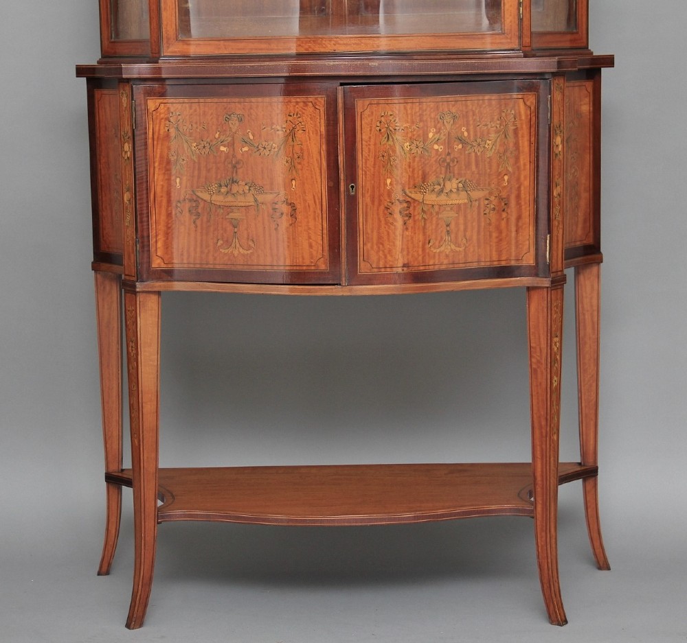 A 19th-century satinwood display cabinet featuring a swan-neck pediment, curved central glass door, and marquetry inlaid lower doors on square tapered legs. View III