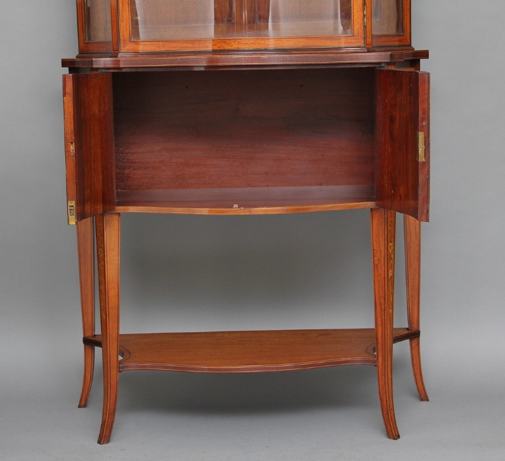 A 19th-century satinwood display cabinet featuring a swan-neck pediment, curved central glass door, and marquetry inlaid lower doors on square tapered legs. View IV