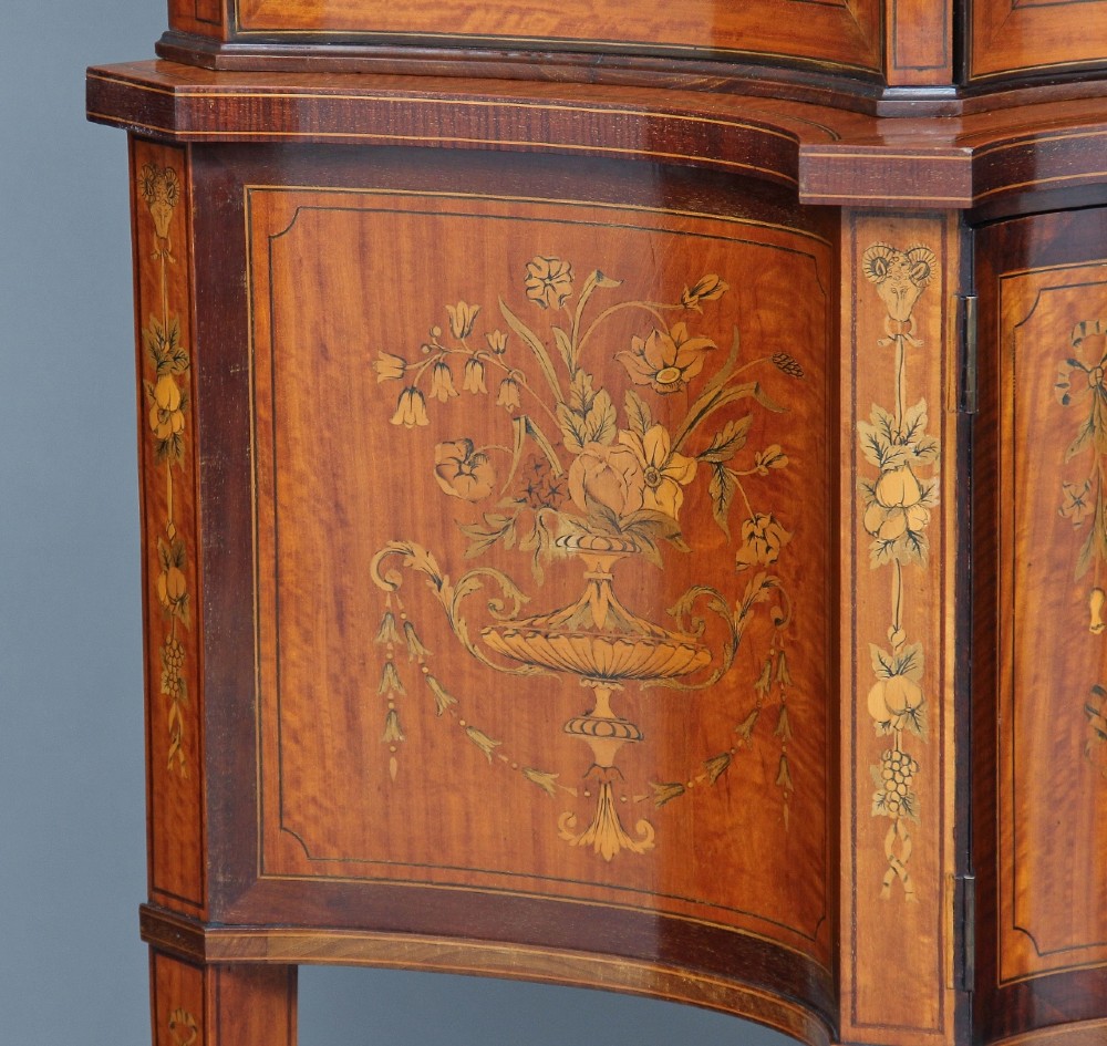 A 19th-century satinwood display cabinet featuring a swan-neck pediment, curved central glass door, and marquetry inlaid lower doors on square tapered legs. View X