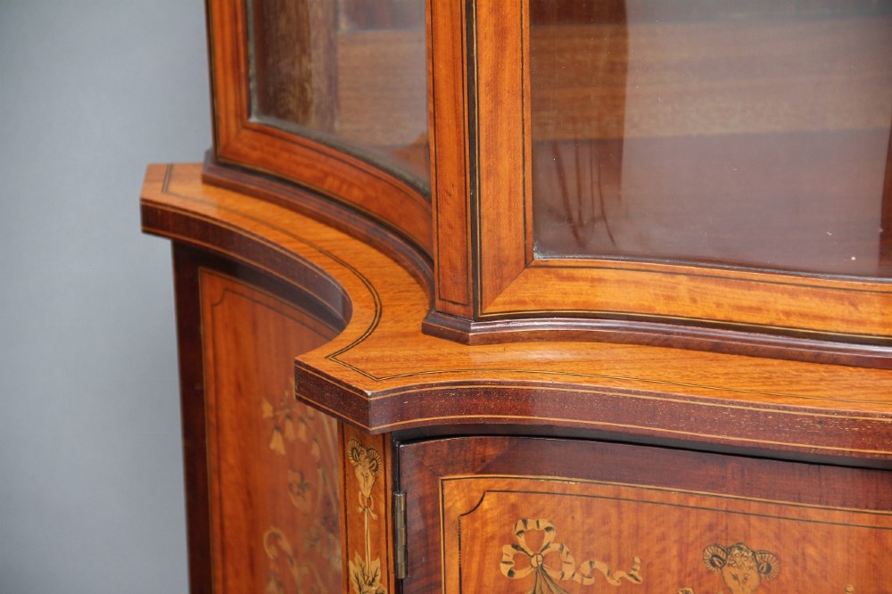 A 19th-century satinwood display cabinet featuring a swan-neck pediment, curved central glass door, and marquetry inlaid lower doors on square tapered legs. View XII