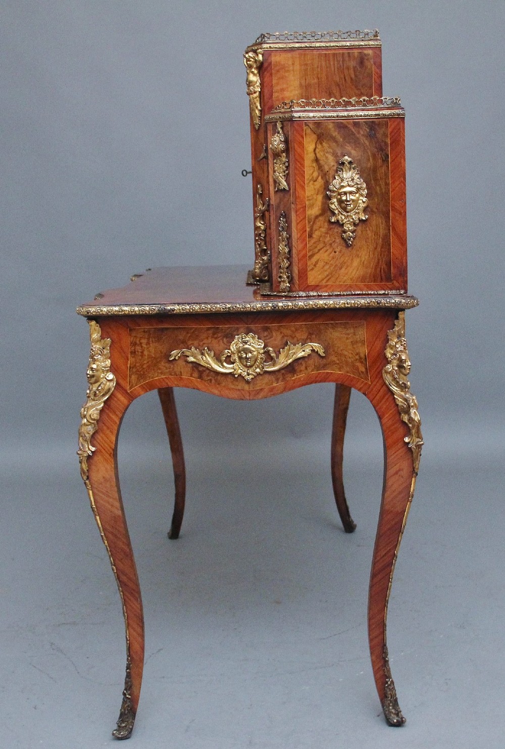 A 19th-century walnut writing desk featuring a raised superstructure with a central glazed cupboard flanked by small drawers, trimmed with gilt-bronze mounts on cabriole legs. View XI