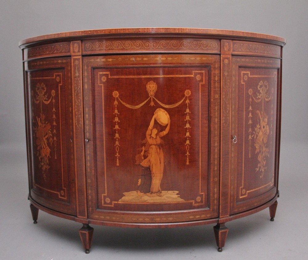 A 19th-century bow-fronted mahogany side cabinet featuring detailed satinwood marquetry panels with classical figures and swags on turned tapered legs.