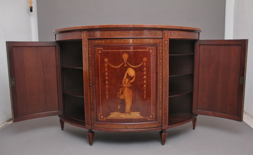 A 19th-century bow-fronted mahogany side cabinet featuring detailed satinwood marquetry panels with classical figures and swags on turned tapered legs. View III