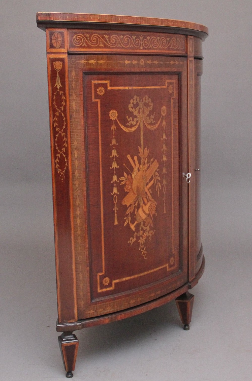 A 19th-century bow-fronted mahogany side cabinet featuring detailed satinwood marquetry panels with classical figures and swags on turned tapered legs. View VI